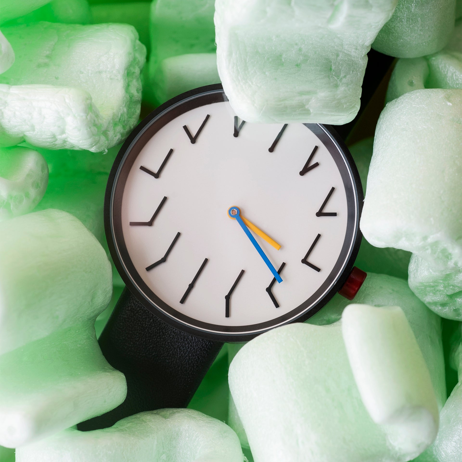 The trio of time - Redundant watch with a minimalist white dial, black strap, and distinctive blue and yellow hands, nestled among soft green packing foam