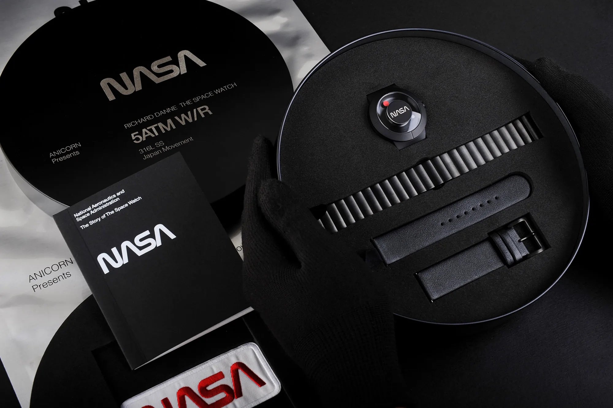 Hands in black gloves presenting The Space Watch by Richard Danne collection with branding book and additional straps on a marbled surface.